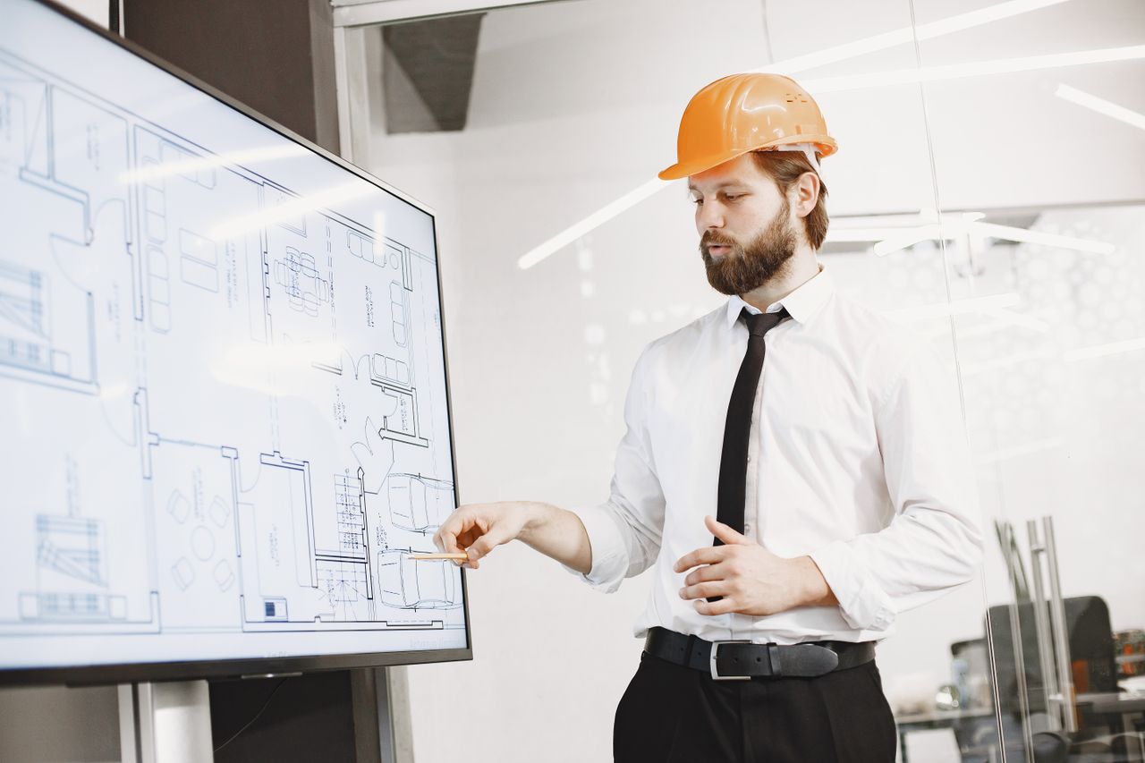 Services Architect in hard hat reviewing blueprints on a screen, discussing plans in modern office.
